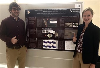 Male and female student (both in khakis and a maroon dress shirt) giving thumbs up. They are standing in front of poster board tacked to the wall.