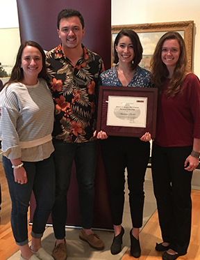 Four students (three girls and one boy) is casual clothing. One of the female students is holding a framed certificate.