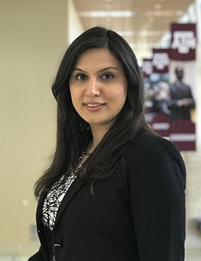 Female student with shoulder length black hair smiling. She is is a black blazer and a black and white shirt with a graphic print on it. Behind her three banners with the Texas A&M logo and an image can be seen in the background.