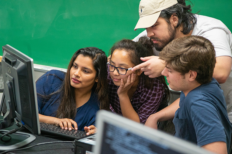 Four students (2 females, 2 males) looking at computer monitor. Three are sitting down, one is standing and pointing at the screen. 