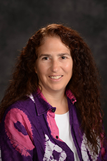 Smiling woman with long, curly brown hair wearing a purple, pink and white button down shirt with white shirt underneath. It is a formal headshot.