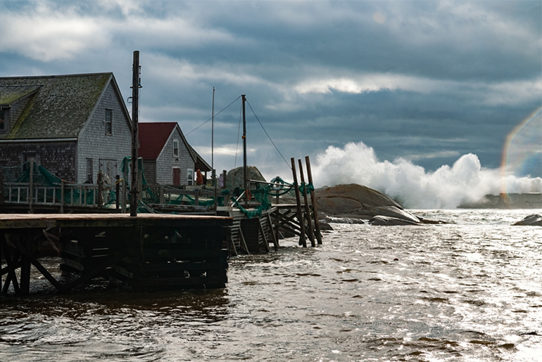 Houses on stilts in a body of water. Part of the dock has broken off and is in the water. In the background there are some rocks and the sky is a dark blue. A little bit of sunshine can be seen reflected off of the water.