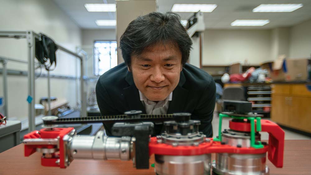 Man in black suit jacket standing behind a table looking down at red and silver prosthetic device.