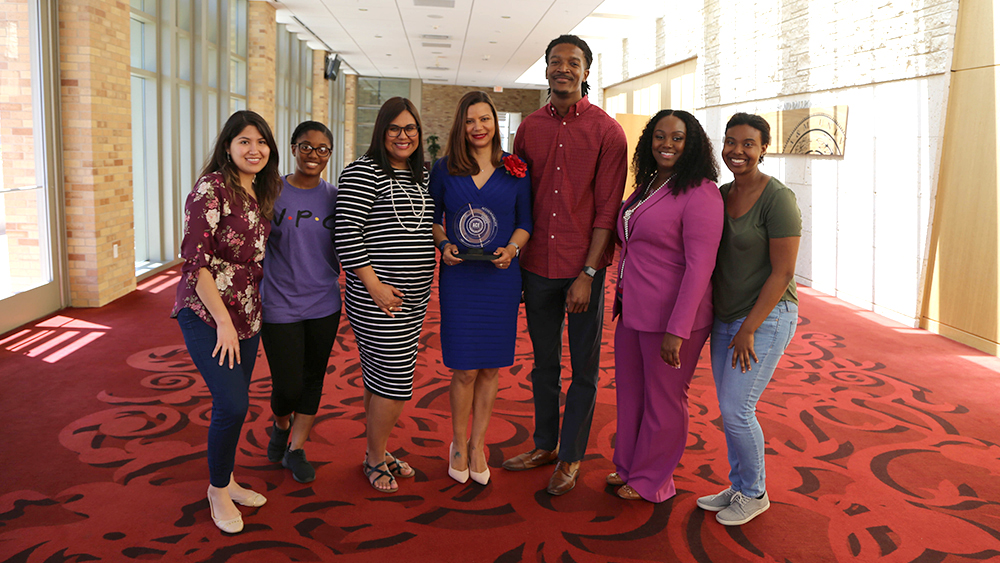 Seven people pose for a photo and a woman in the middle holds an award