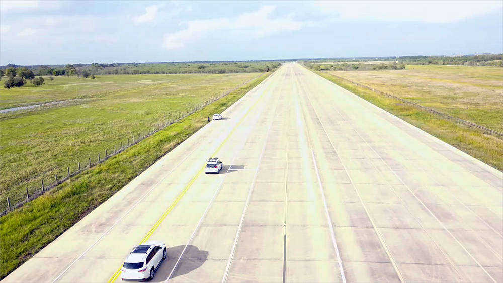 An autonomous vehicle driving down an open testing road site followed by a car driven by a human.