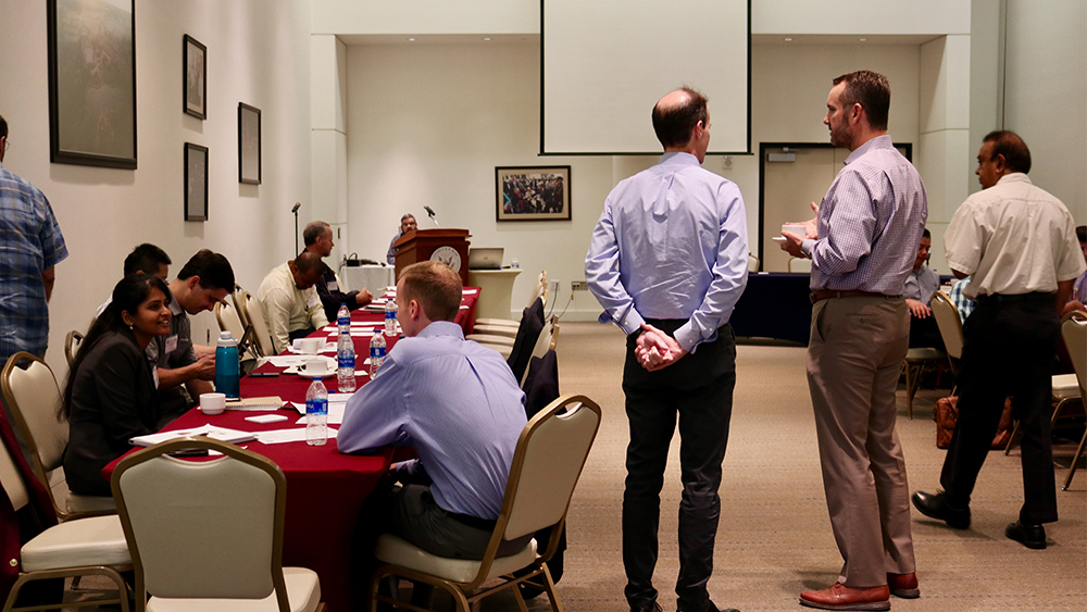 Two individuals standing and speaker. Other individuals sitting and speaking. 