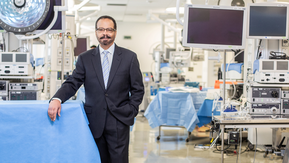 Dr. Roderic I. Pettigrew, wearing a dark suit, white shirt, striped light blue tie, and glasses, stands in a room filled with medical equipment. He has one hand in his pocket, and another hand resting on a table covered by a blue sheet.