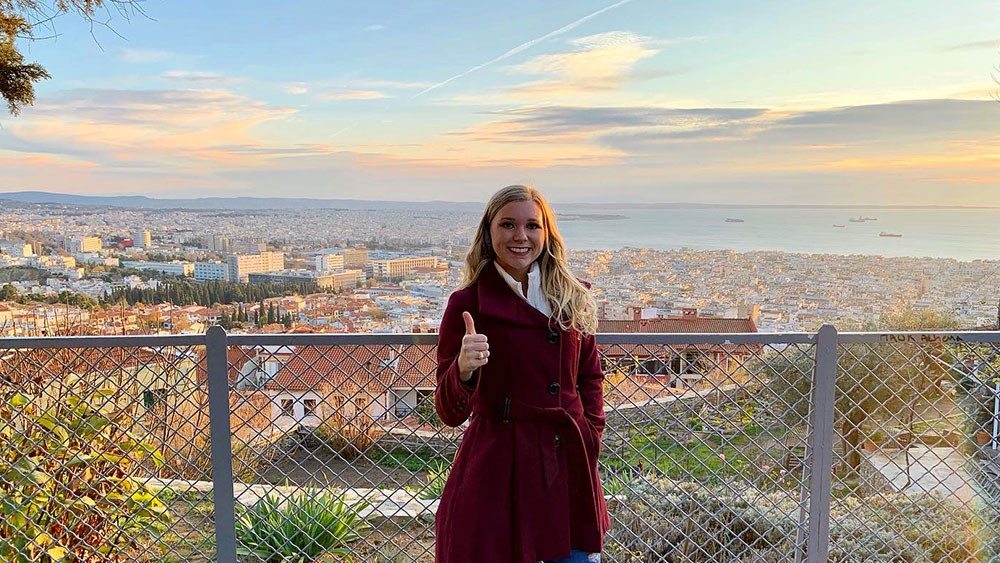 Lauren Williams stands in front of a scenic overlook of a sprawling city. She is wearing a maroon coat and holding up a gig 'em hand sign.