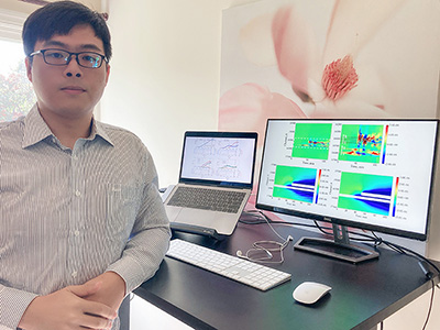 Man standing beside a tall desk where a displayed laptop and additional screen show several graphs of lines and colored areas