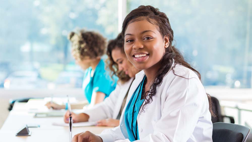 Three medical students write while sitting at a table. One of them is looking at the camera and smiling