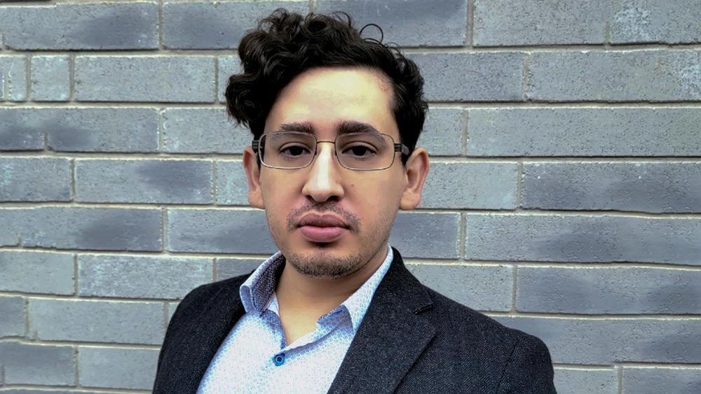 a man in a blue button down shirt and a black blazer gazes at the camera for a headshot. he is wearing glasses. 