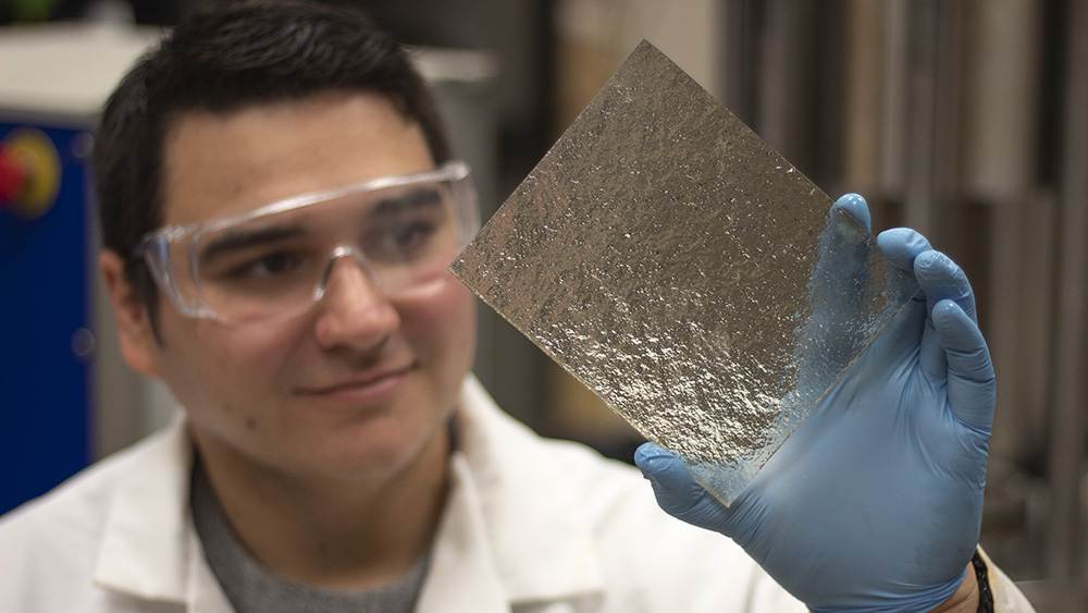 Male student in lab coat and safety glasses holding and studying clear resin 3D printout in his gloved hand