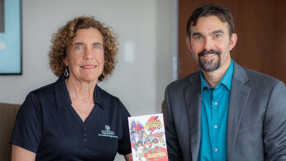 Two faculty members sit at a table. One holds a small booklet. Both are smiling at the camera.