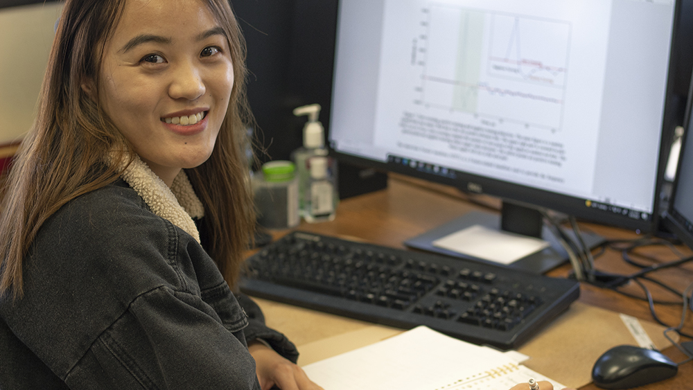 female graduate student seated at desk while using computer and writing notes in lab journal