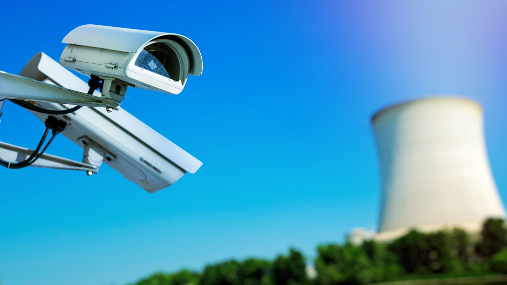 Two security cameras monitor a nuclear power reactor site located behind a body of water and shrubbery. 