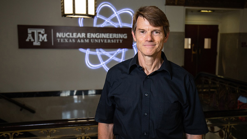 Dr. Marvin L. Adams standing in a room with the Department of Nuclear Engineering and  a nuclear fission symbol in the background.