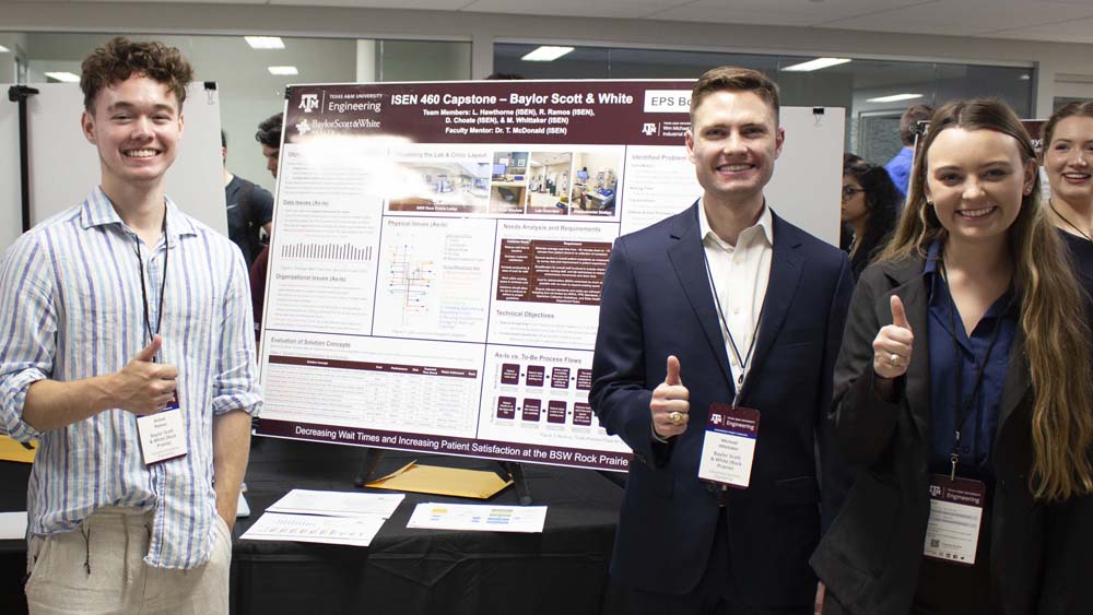 Three students in business formal stand in front of a poster. They are all smiling at the camera.