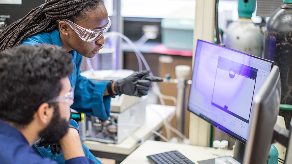 Female and male graduate students in lab and dressed in safety gear are discussing images on a computer screen