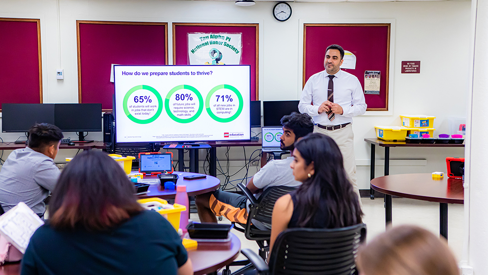 Professor presents data on a flatscreen TV to workshop participants sitting at tables. Information displayed: How do we prepare students to thrive?