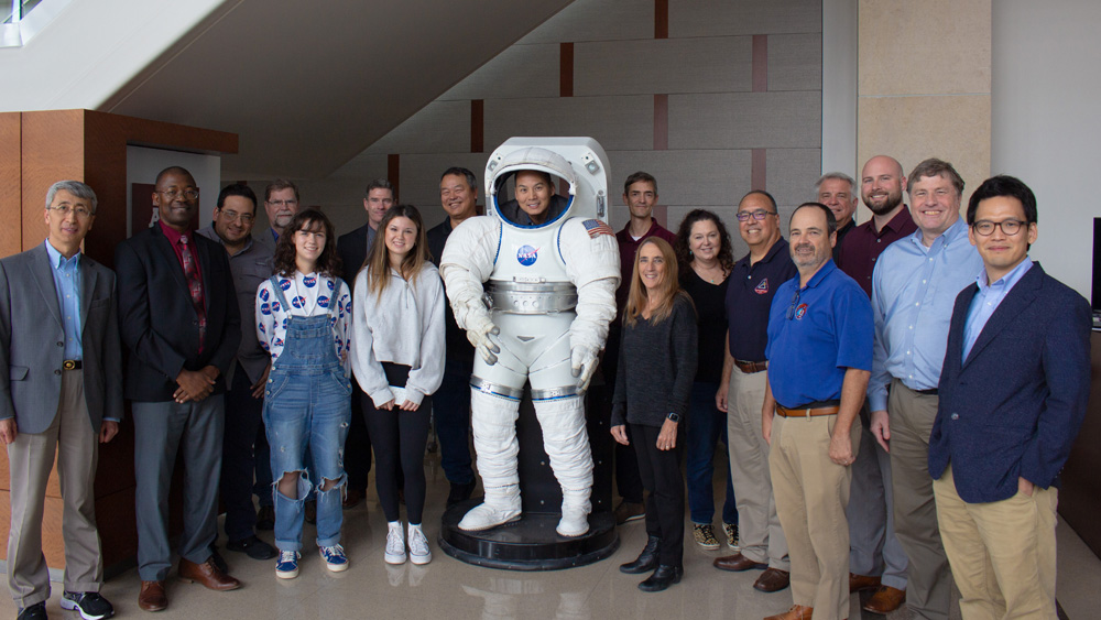 A group of NASA representatives, their families and Texas A&amp;M faculty stand together around a life-sized space suit model and smile at the camera.