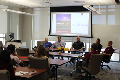 Five men sit at a table, talking with a room of students and faculty. A NASA Day informational slide is on a screen behind the men.