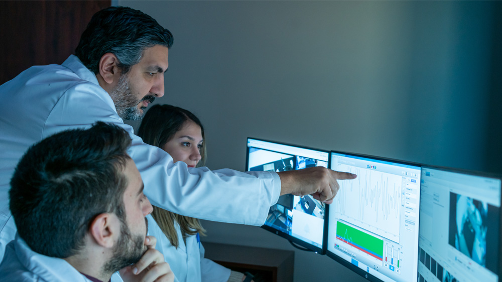 A faculty member and two students sit in front of three computer screens. The faculty member is pointing at the middle screen, where data points are connected through jagged up and down lines.