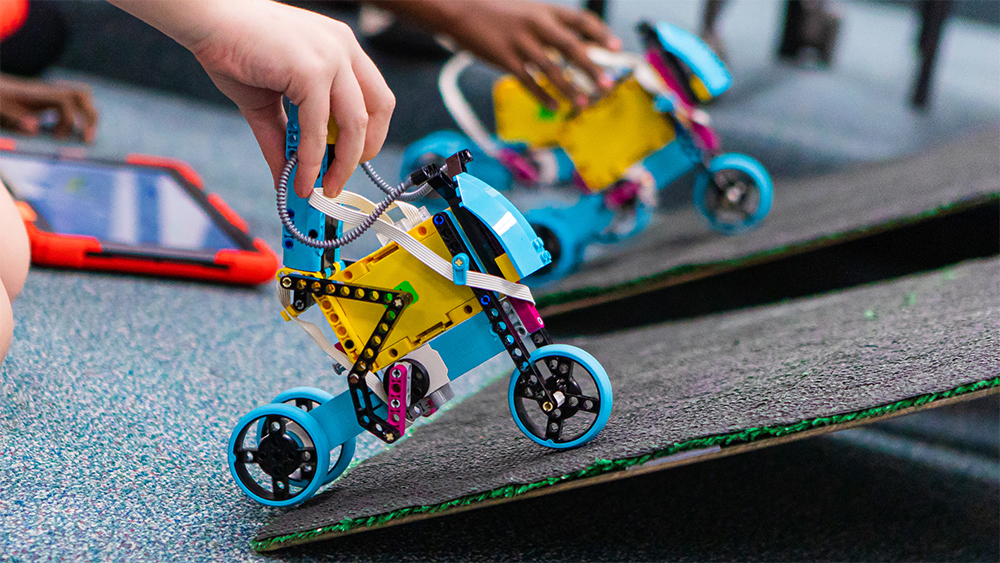 Students place their colorful wheeled robots on cardboard ramps and there is a red tablet on the ground between the students.