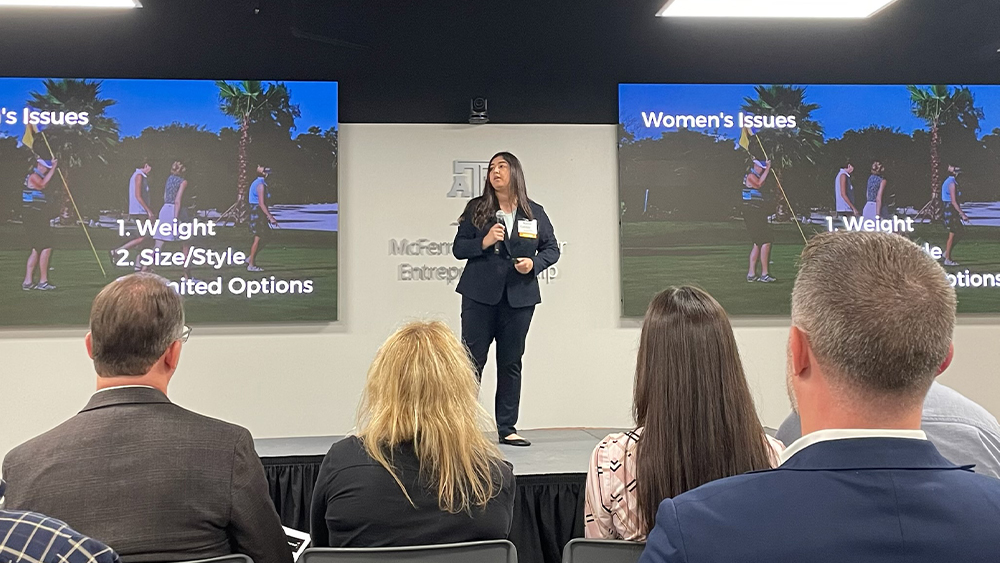 Katie Calderon presents at the Aggie PITCH event in front of a crowd. In the background, a screen reads: Women’s issues, 1. Weight, 2. Size/style, 3. Limited options.