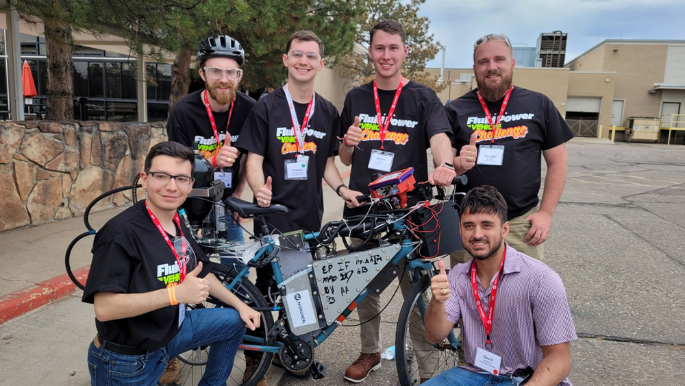 The team stands and kneels around their bike while they give a thumbs up to the camera.