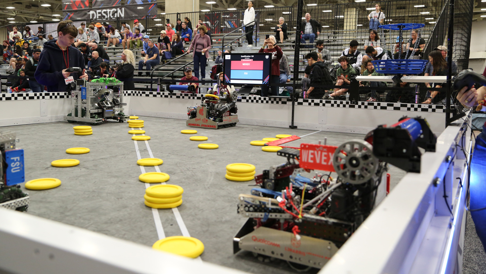 An arena with multiple yellow discs and robots on the edges of the arena trying to pick up the discs to shoot them into high and low goals.