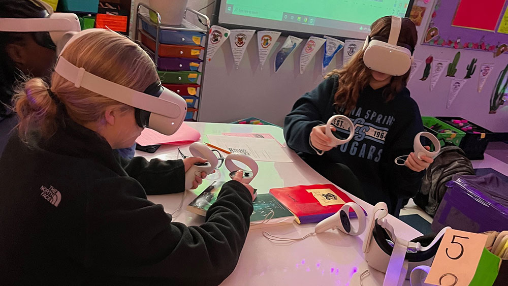 Two students sit at a classroom desk playing with virtual reality headsets and controllers.