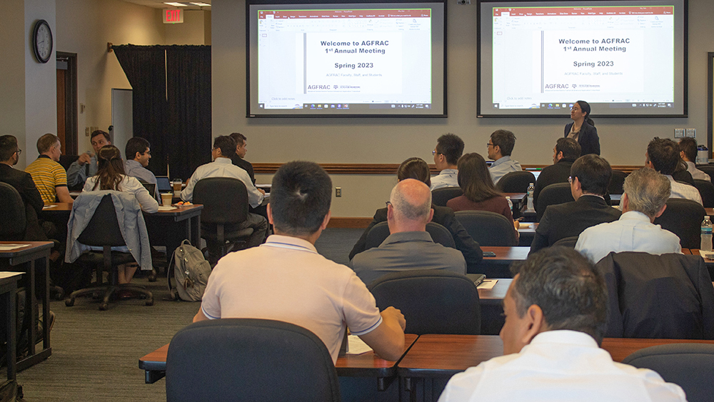 Several people seated at rows of tables in a large room where Dr. Kan Wu is speaking in front of two projection screens on the wall behind her.