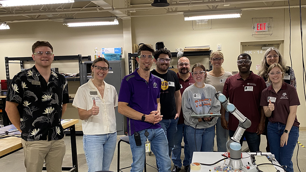 Ten people smiling at the camera stand behind a worktable with a robot arm located in a workshop. Two people are giving thumbs-up hand gestures. 