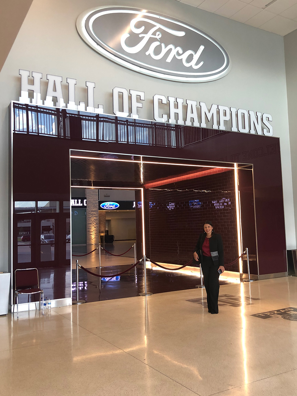 Dr. Jennifer Carvajal is standing outside of the entrance of the Hall of Champions at Texas A&amp;M in front of the rope, wearing black pants, a black jacket, and a maroon shirt. 