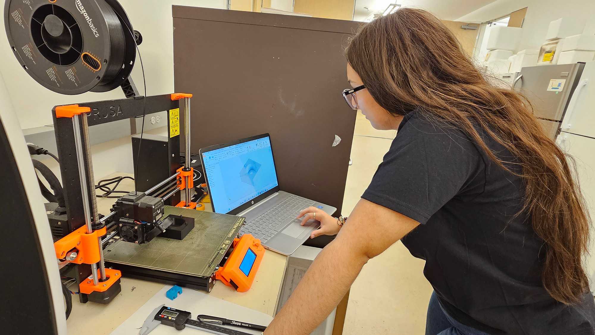 Girl in black shirt stands in front of laptop computer and table top 3D printer working on printed project.