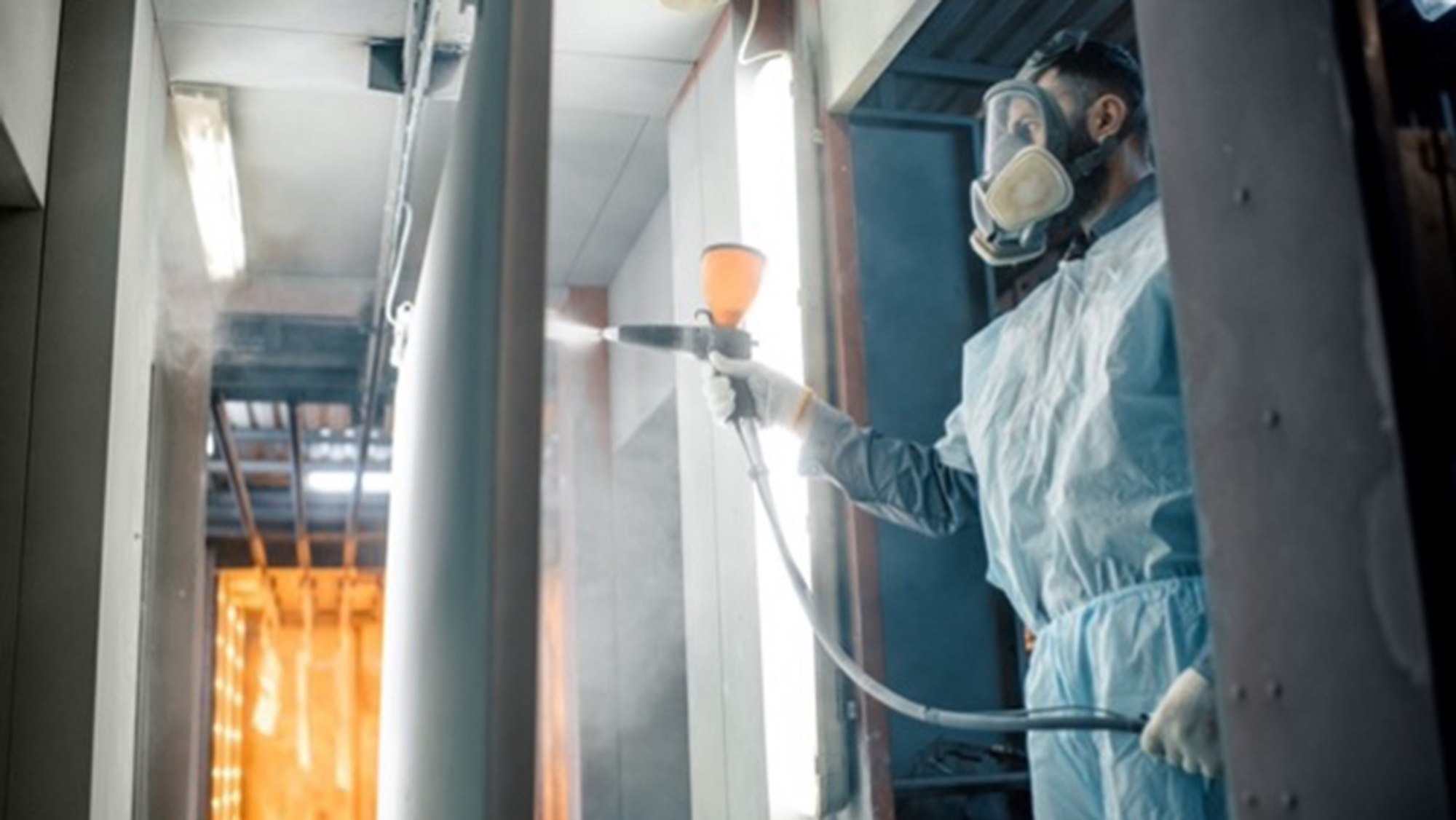 A man in a protective suit wearing a gas mask is spraying a sheet of metal with a layer of protective covering with a machine in a lab.