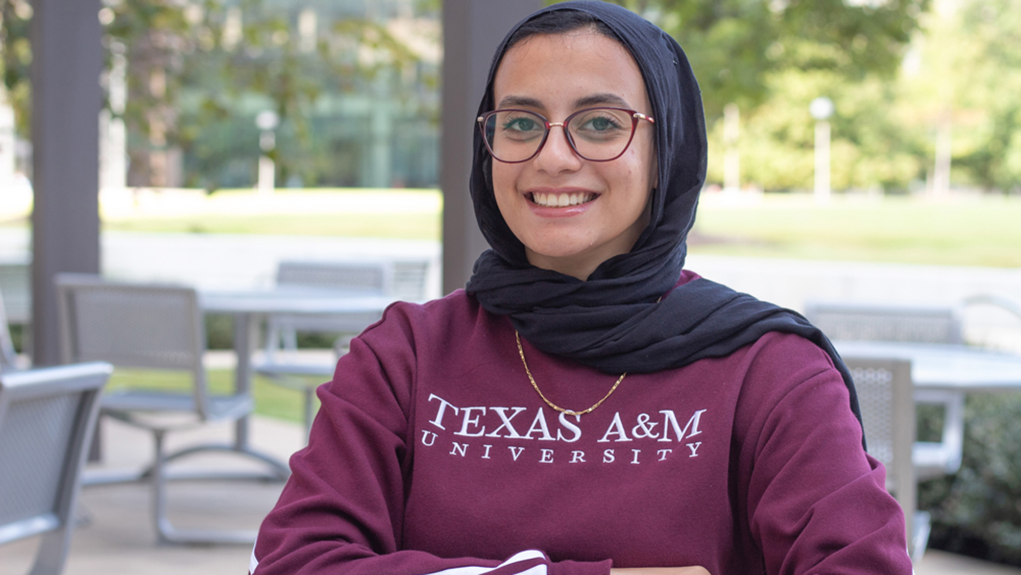 Smiling female student sitting outdoors wearing glasses, a hijab and a long-sleeve shirt.