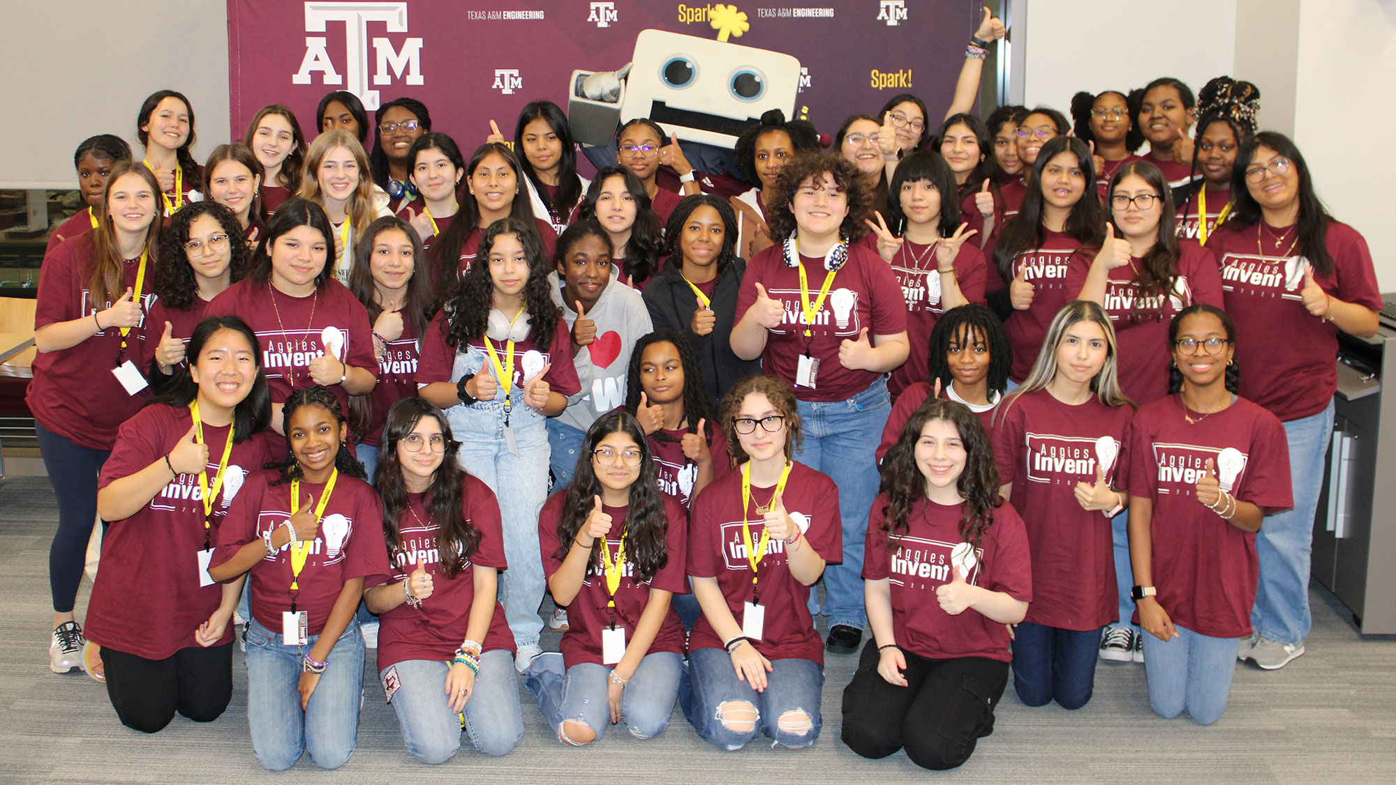 A group of over 30 high school female students wearing maroon t-shirts stand in front of a maroon backdrop along with Spark-e the robot.