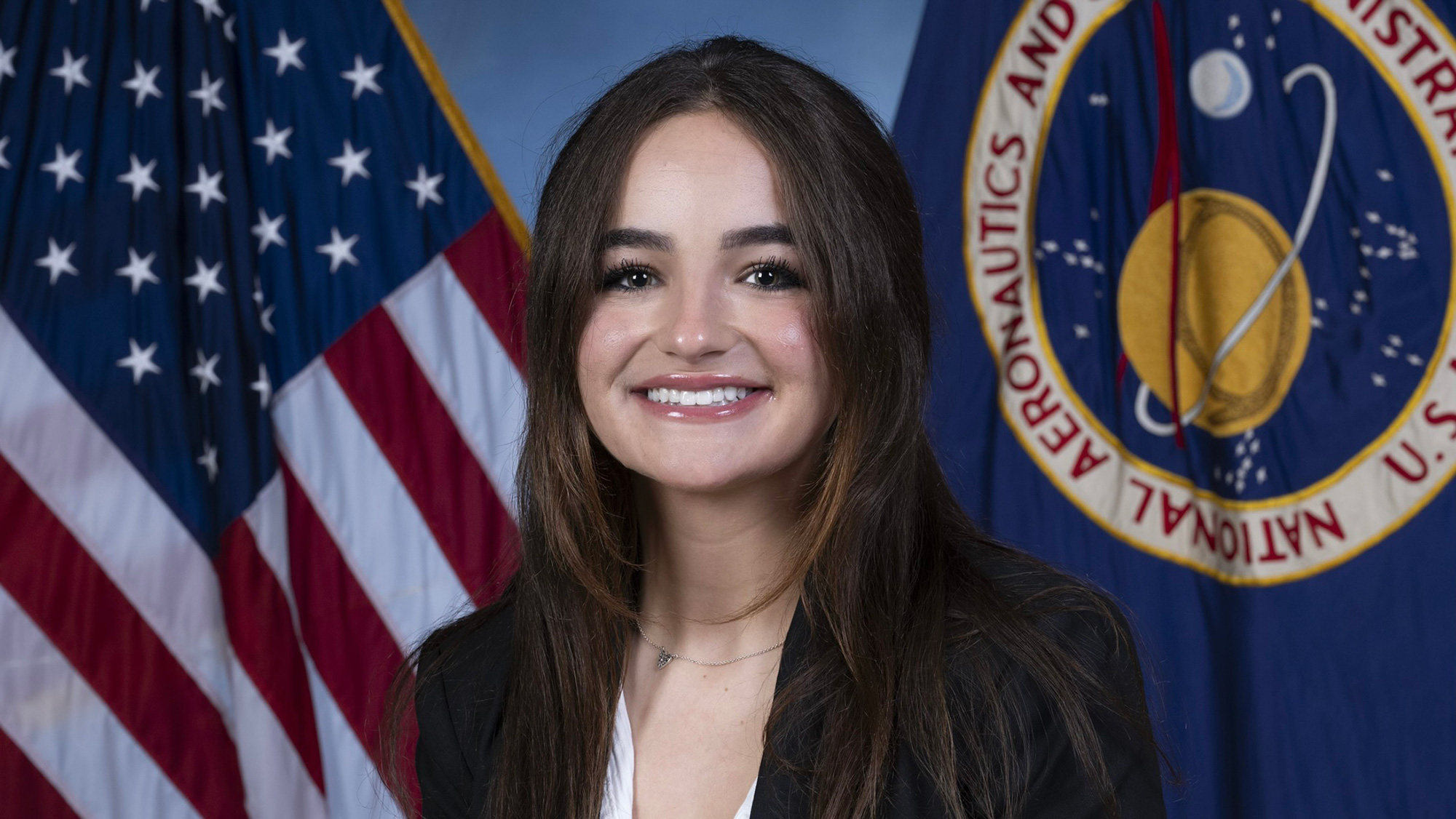 Sofi English, a young woman with long dark hair sits in front of American flag and NASA flag in the background.