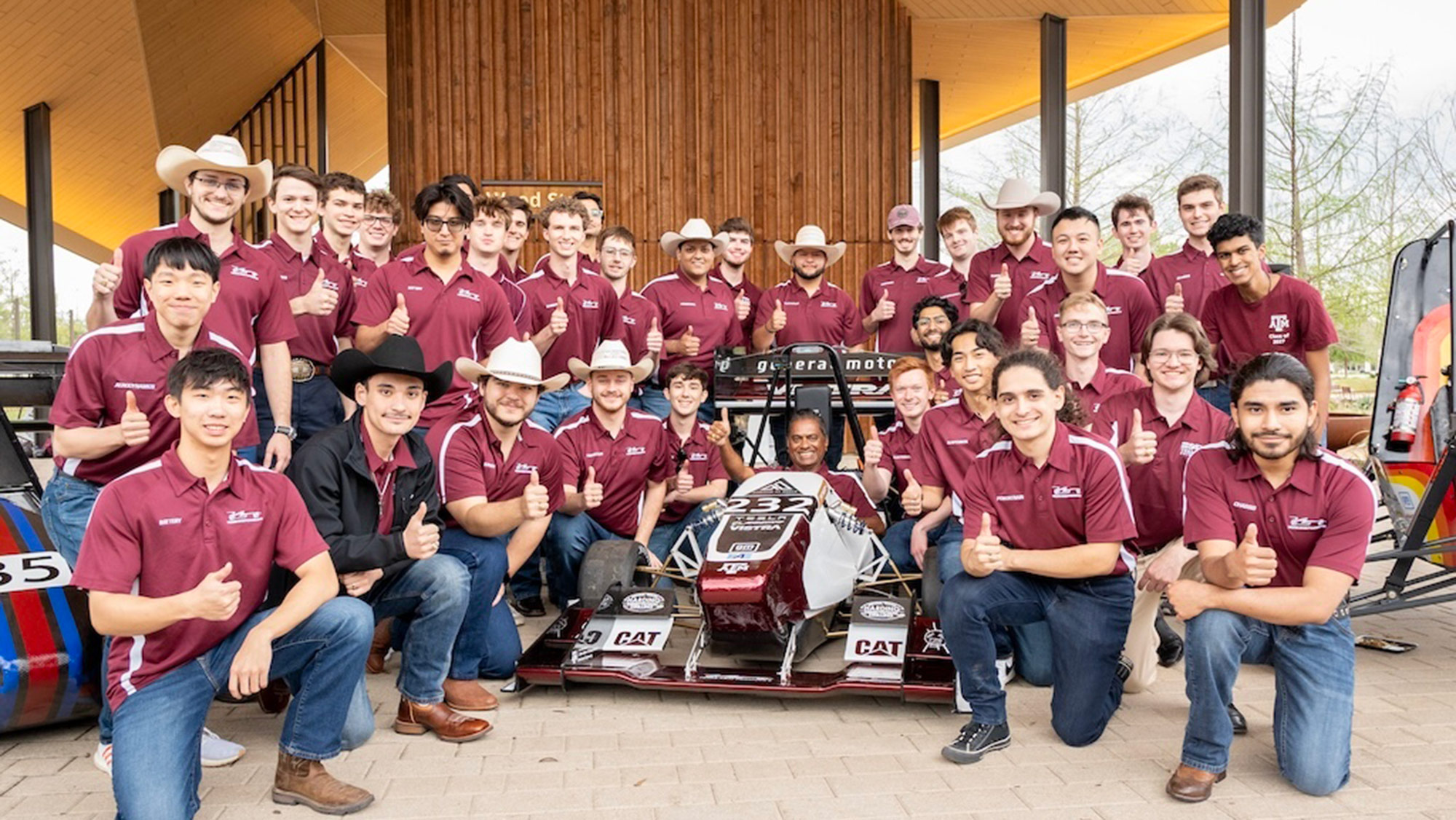 A group of students and a faculty member posing with a formula-style race car under a wooden pavilion.