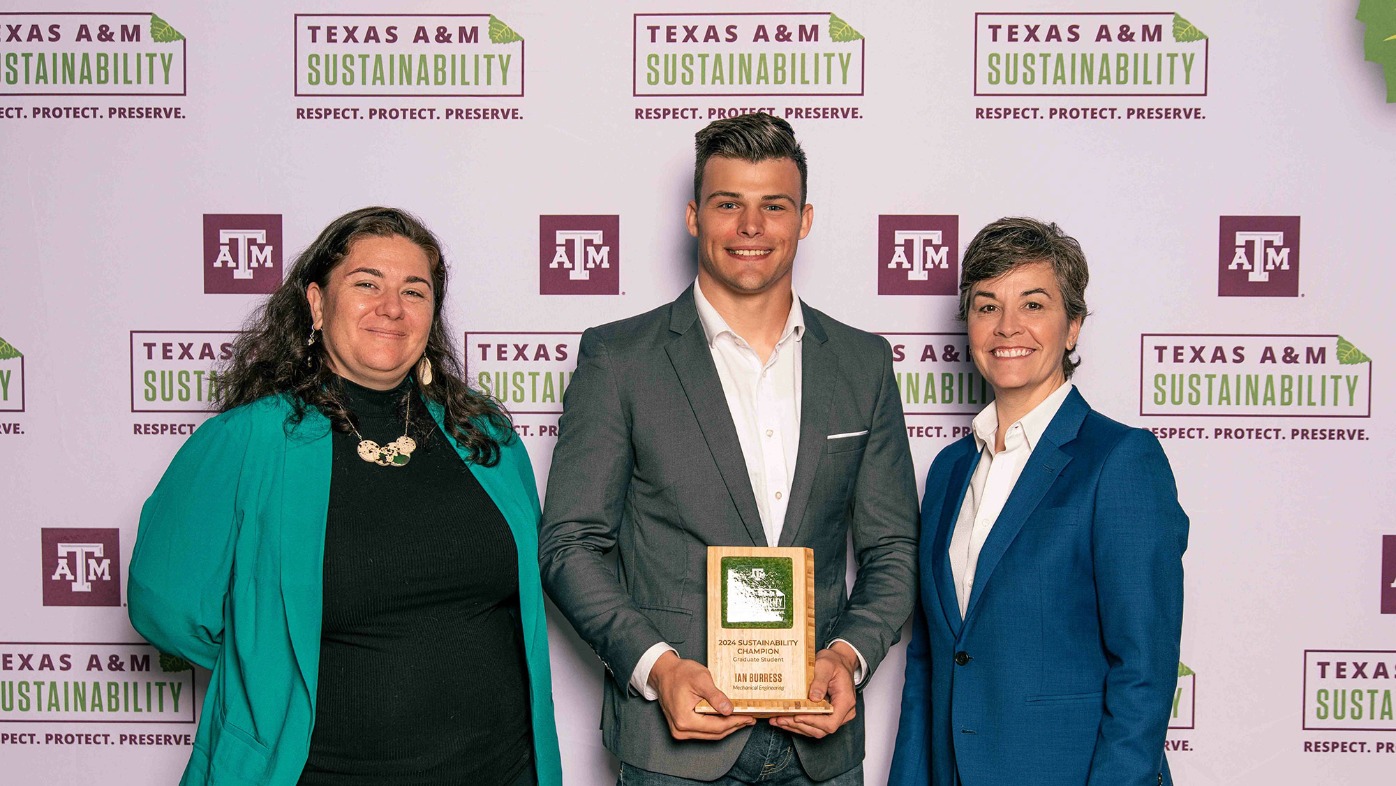 A man holds a plaque and stands by two women against a background that says Texas A&amp;M Sustainability. Respect. Protect. Preserve.