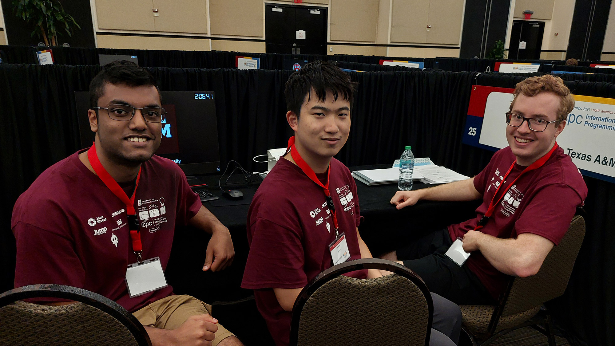 Three men in matching maroon shirts sitting at a computer.