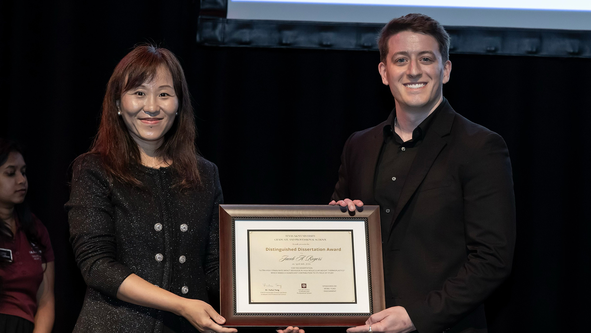 Two people smiling while holding an award plaque on stage.