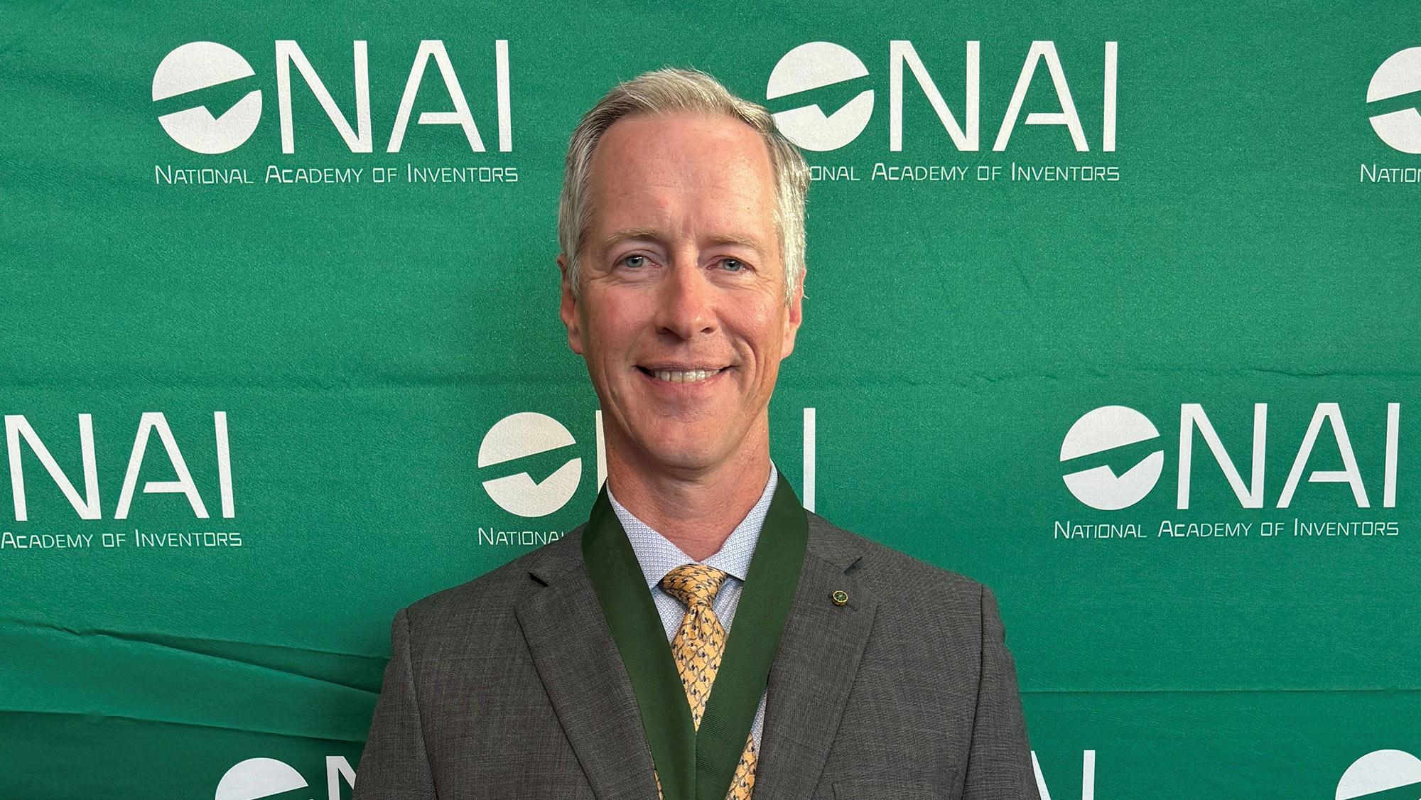 A man is wearing a medal in front of a banner with the National Academy of Inventors logo.