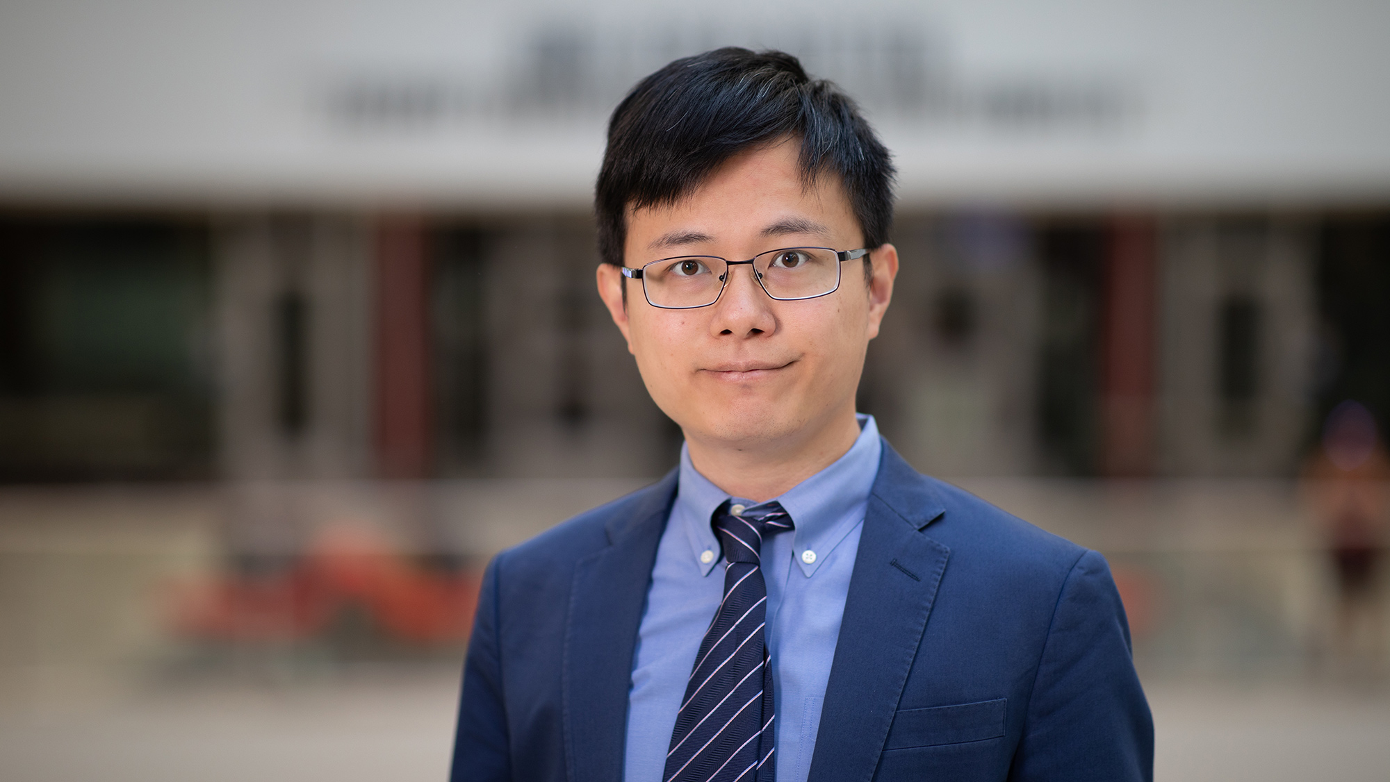 A man with glasses in a blue suit in front of a blurred background. 