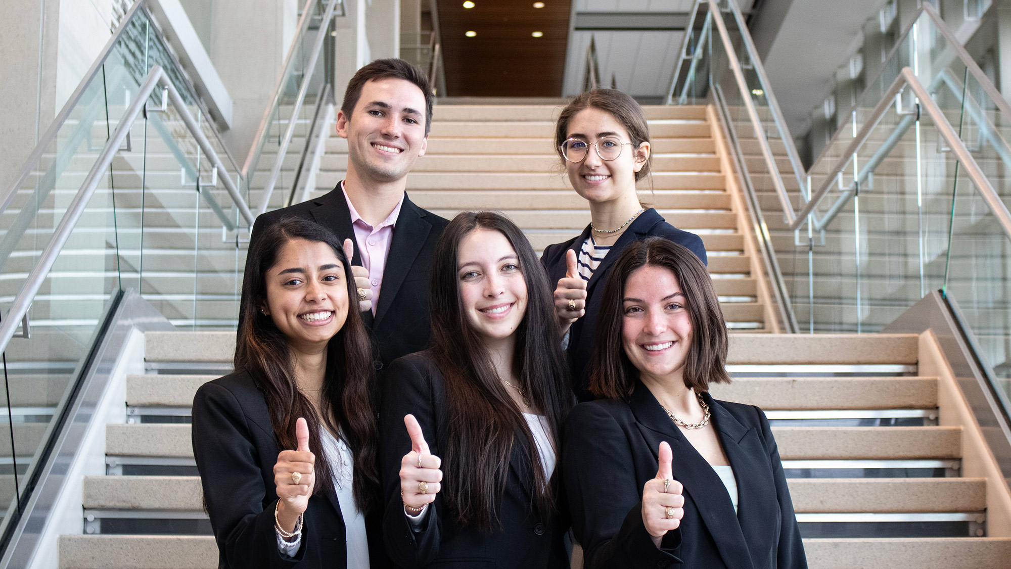 Five students dressed in professional attire with their thumbs up on the steps of a campus building.