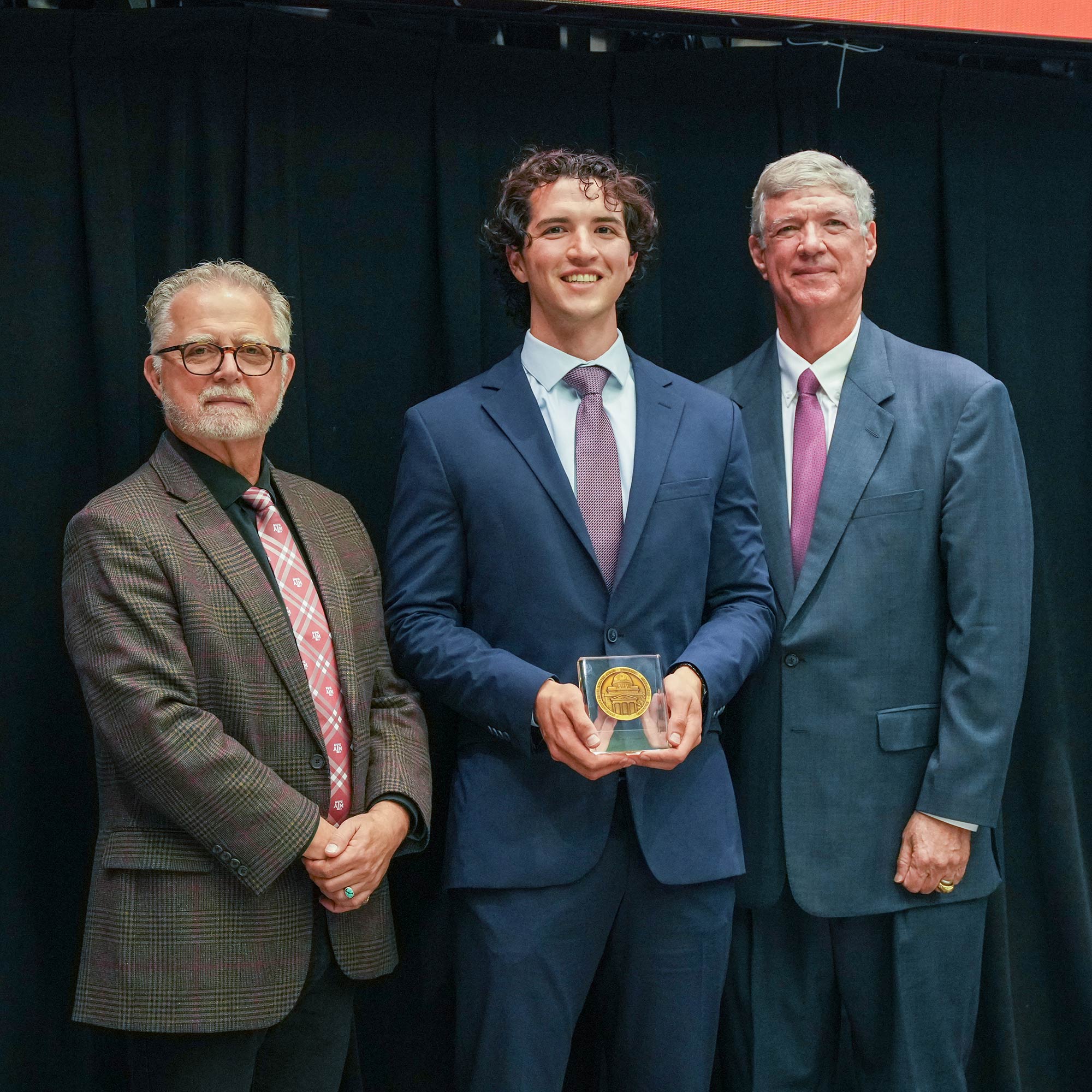 Three men standing, one with an award.