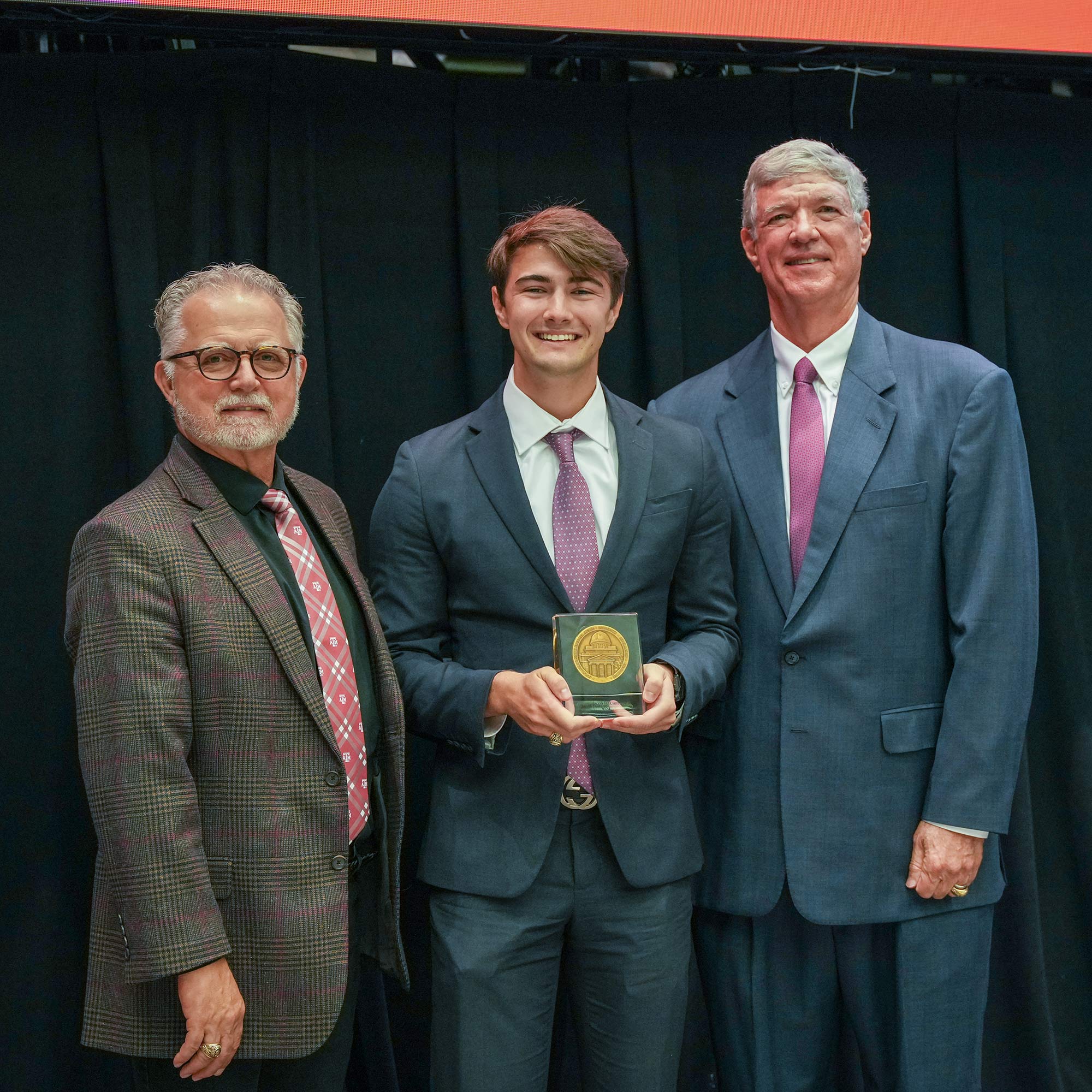 Three men standing, one with an award.