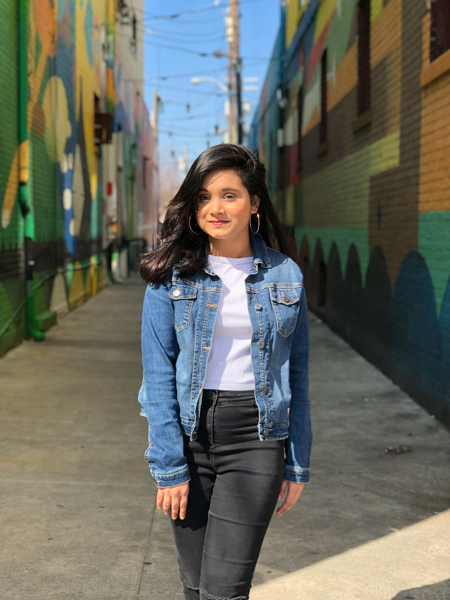 A young woman standing in a colorful alleyway wearing a denim jacket over a white shirt and black jeans.  