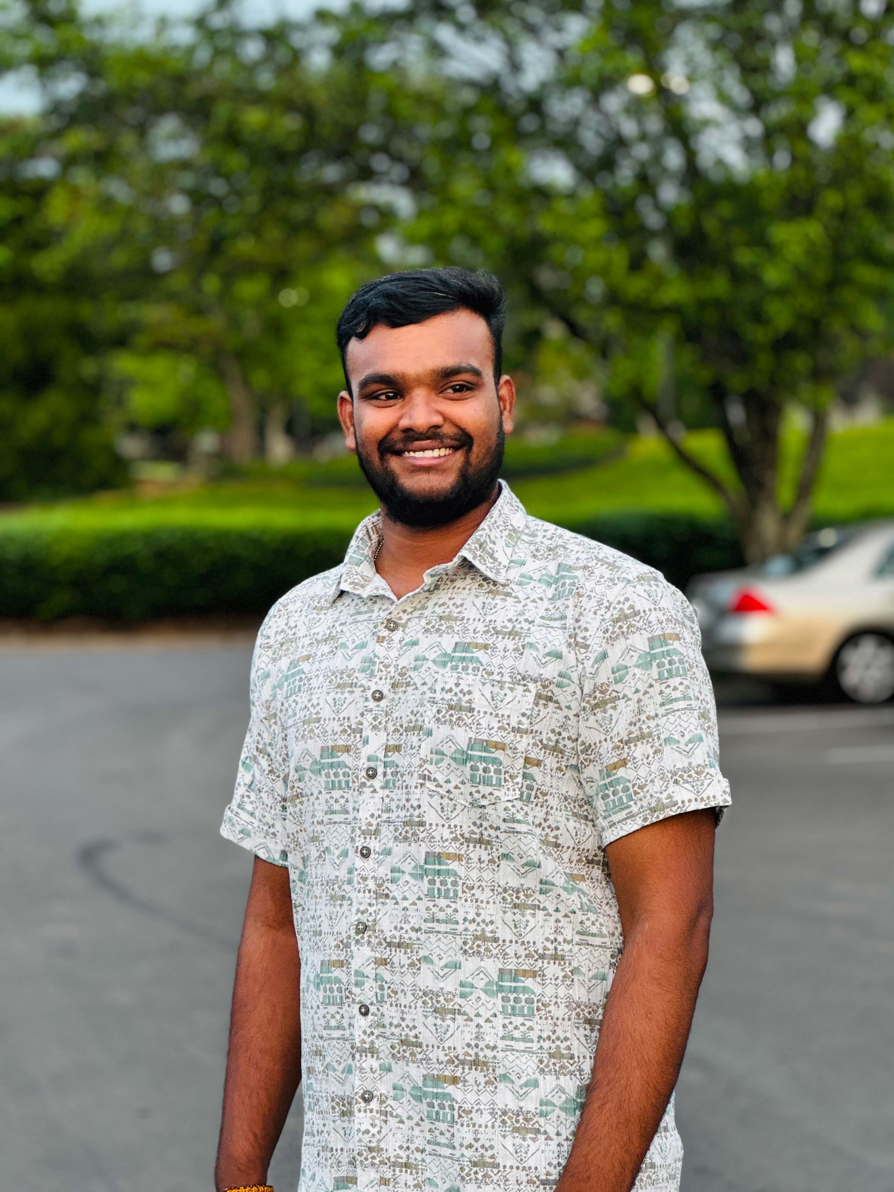 A young man stands outdoors wearing a patterned short-sleeve shirt.